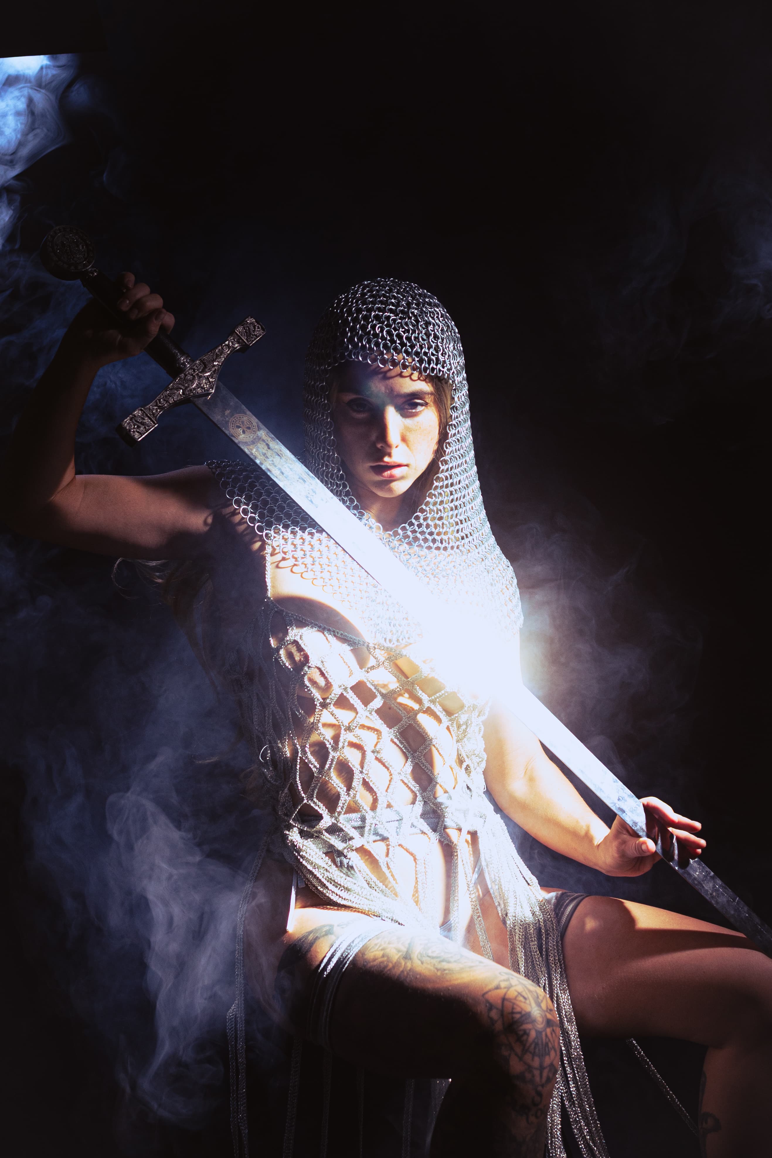 Figure in a chainmail hood holding a sword through haze.