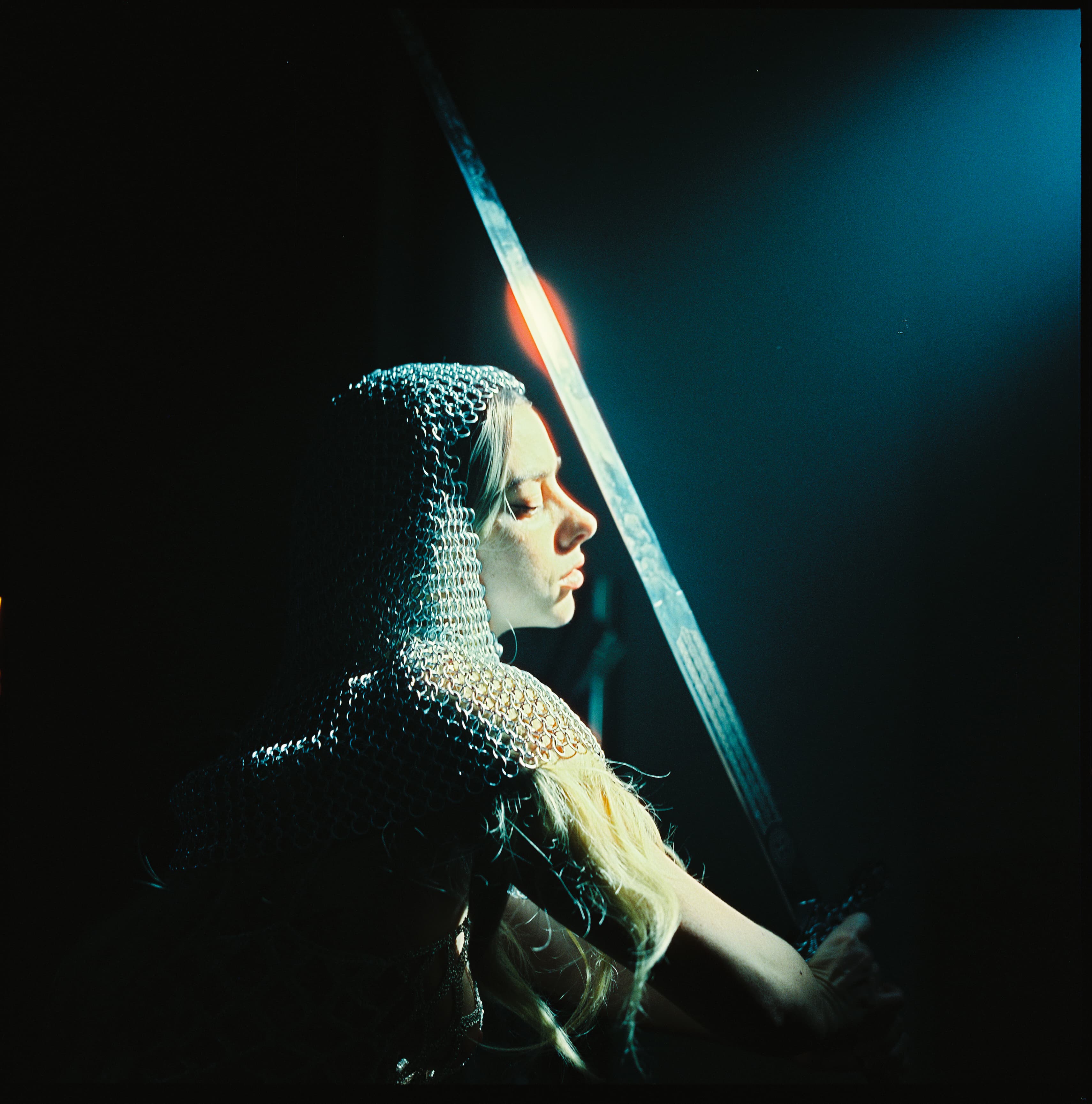 Figure in a chainmail hood beside a beam of teal light.