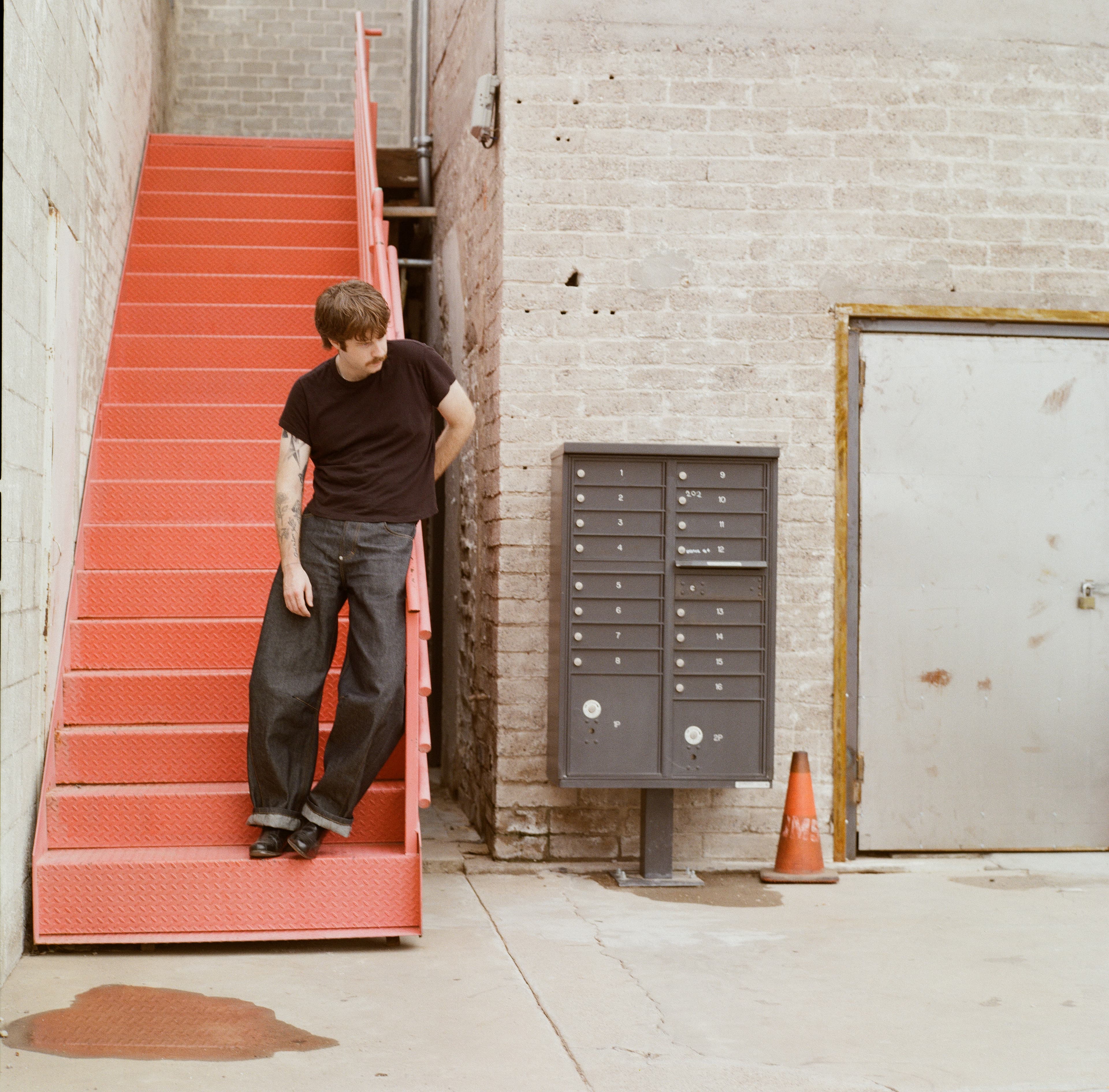 Man leaning on a red staircase beside a brick wall and mailboxes.