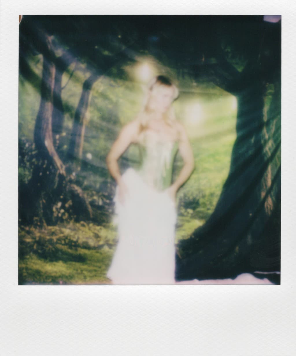 Polaroid of a woman in a corset and white skirt in front of a forest backdrop.