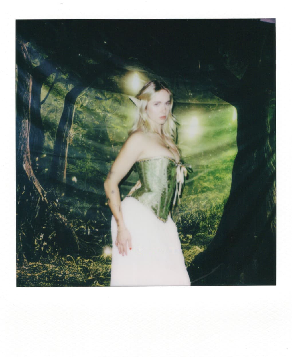 Polaroid of a woman in a green corset turning toward the camera in a forest set.