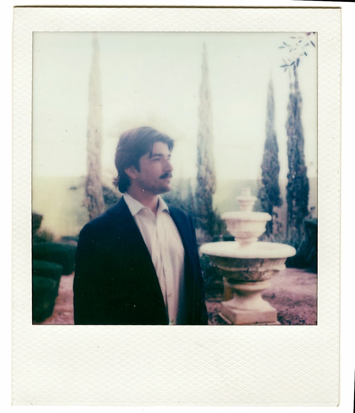 Polaroid of a man in a dark jacket beside a garden fountain.